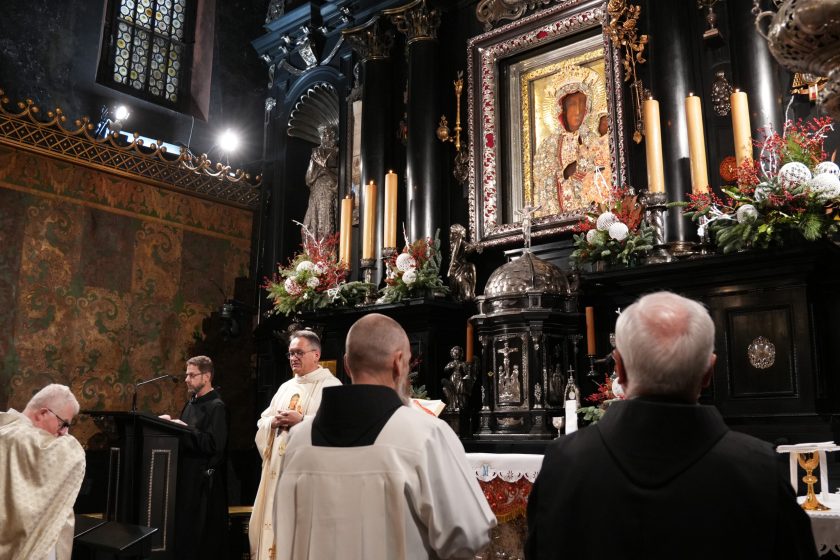 Szeroki kadr z wnętrza kaplicy na Jasnej Górze: czarny ołtarz z licznymi świecami i dekoracjami roślinnymi oraz obraz Matki Bożej Częstochowskiej w ozdobnej ramie. Na dole kadru widoczni uczestnicy liturgii: duchowni w jasnych szatach i zakonnicy, część odwrócona tyłem do aparatu. Z lewej strony przy pulpicie stoi zakonnik w czarnym habicie, obok kapłan w białych szatach; w tle wysoka, bogato zdobiona ściana i witrażowe okno