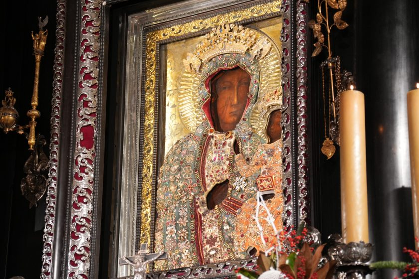 Zbliżenie na obraz Matki Bożej Częstochowskiej w bogatej, srebrno-złotej ramie; postacie Matki Bożej i Dzieciątka ubrane w ozdobne, wysadzane kamieniami sukienki i korony. Po prawej stronie kadru stoją wysokie świece o jasnym kolorze. U dołu widoczny fragment dekoracji roślinnej z czerwonymi akcentami