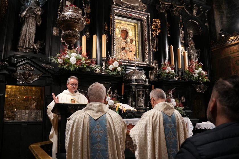 Kadr na ołtarz w kaplicy: w centrum obraz Matki Bożej Częstochowskiej, poniżej tabernakulum i elementy srebrnej ornamentyki. Na pierwszym planie od tyłu stoją dwaj duchowni w jasnych ornatach z niebieskimi pasami zdobionymi krzyżami. Po lewej kapłan w białych szatach przemawia przy ambonie, z uniesioną dłonią; w tle ciemne kolumny, świeczniki i bogate zdobienia ołtarza