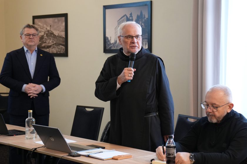 Sala konferencyjna: zakonnik w czarnym habicie stoi przy stole i mówi do mikrofonu, zwrócony w stronę uczestników. Po lewej w tle stoi mężczyzna w garniturze z założonymi rękami. Po prawej przy stole siedzi zakonnik w czarnym stroju, notuje w zeszyci; obok laptop, butelka napoju i notatnik. Na ścianie wiszą czarno-białe fotografie w ramkach.