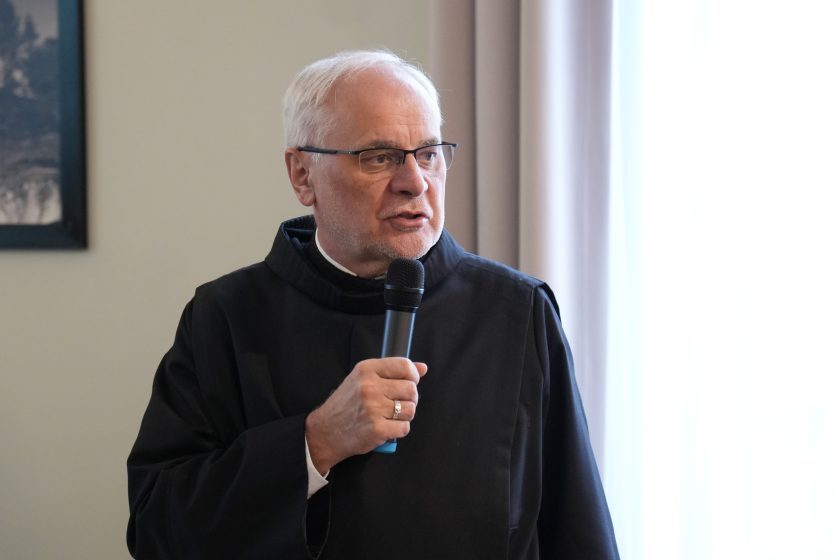 Zbliżenie na zakonnika w czarnym habicie mówiącego do mikrofonu; trzyma mikrofon jedną ręką i patrzy w bok, jak podczas wypowiedzi. W tle jasna ściana oraz fragment zasłony i ramki ze zdjęciem, tło jest lekko rozmyte