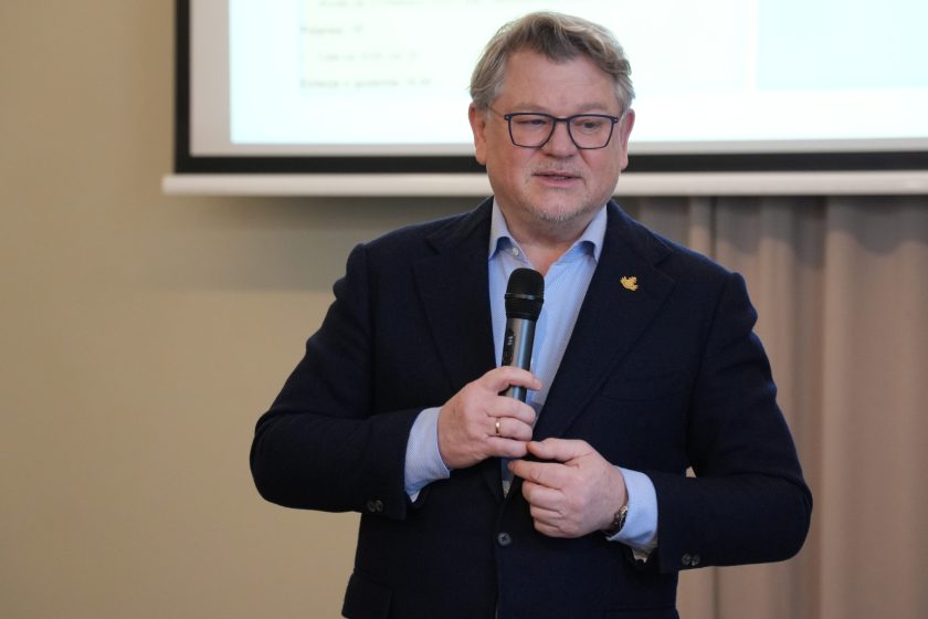 Portret mężczyzny w garniturze i okularach, trzymającego mikrofon na tle ekranu projekcyjnego. Za nim widoczny zarys slajdu prezentacji, a obok zasłona przy oknie; mężczyzna wygląda na prowadzącego część spotkania