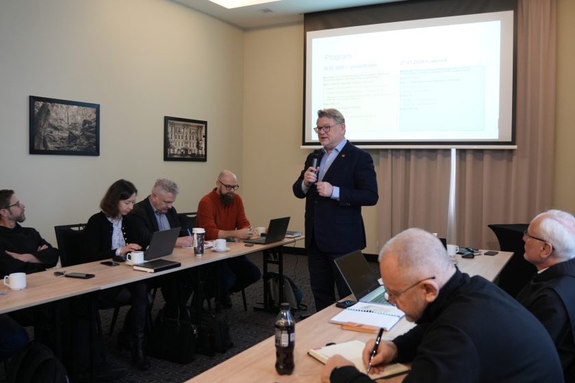 Szeroki plan sali konferencyjnej: prowadzący w garniturze stoi i mówi do mikrofonu przed ekranem z wyświetloną prezentacją (slajd z nagłówkiem i listą punktów programu). Uczestnicy siedzą przy ustawionych w podkowę stołach, pracują na laptopach i robią notatki; na blatach stoją kubki, szklanki oraz notatniki. Na pierwszym planie po prawej zakonnik pochylony nad notesem zapisuje uwagi długopisem