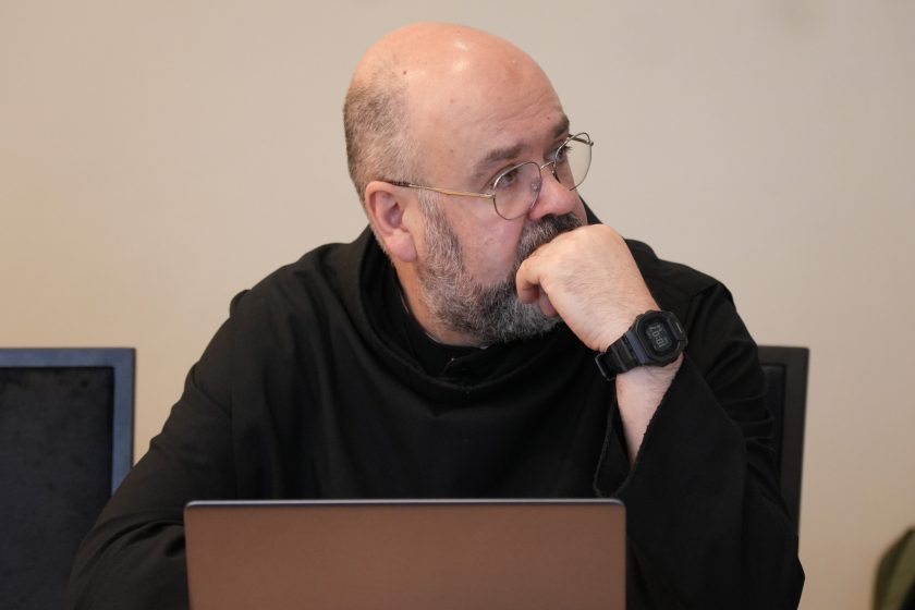 Zbliżenie na zakonnika w czarnym stroju siedzącego przy stole; opiera brodę na dłoni i patrzy w bok z zamyślonym wyrazem twarzy, jak podczas słuchania. Na stole przed nim widoczna krawędź laptopa; na nadgarstku ma zegarek