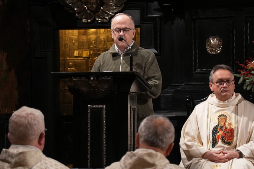 Mężczyzna w świeckim stroju czyta tekst przy ambonie w kaplicy, obok siedzi kapłan w białych szatach liturgicznych. Na pierwszym planie widoczne głowy uczestników nabożeństwa