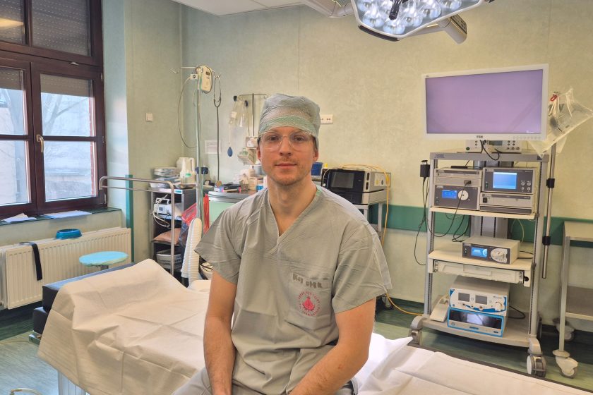 Mężczyzna w stroju operacyjnym siedzi na stole zabiegowym w sali operacyjnej, zwrócony przodem do aparatu. Ma na sobie jasnoszary strój chirurgiczny i czepek, a na twarzy neutralny, skupiony wyraz. W tle widoczna jest aparatura medyczna, monitor, wózek z urządzeniami oraz elementy wyposażenia sali operacyjnej. Nad stołem zamontowana jest lampa operacyjna, a przez okno wpada światło dzienne