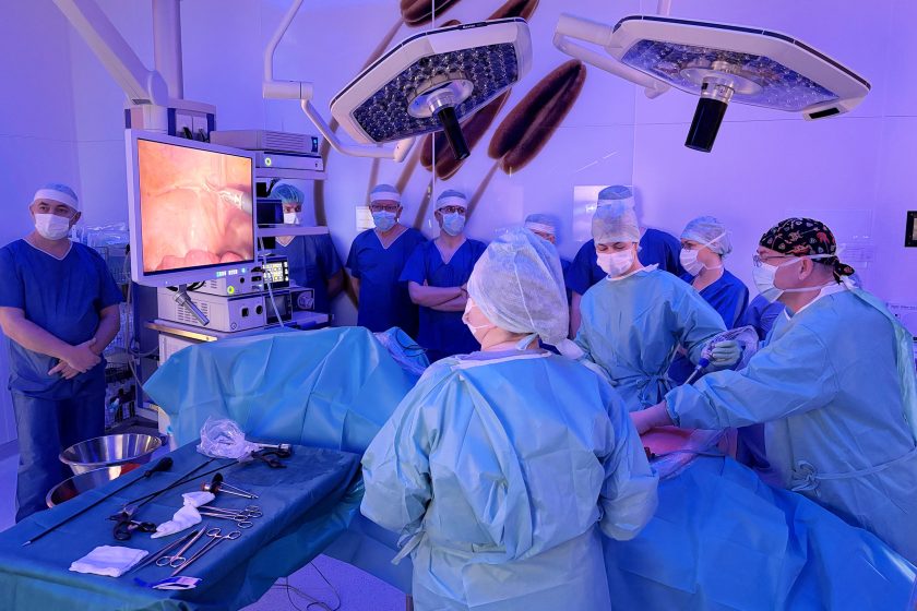 W nowoczesnej sali operacyjnej lekarze i rezydenci w odzieży ochronnej uczestniczą w zabiegu laparoskopowym. Część zespołu obserwuje przebieg operacji na dużym monitorze medycznym, na którym wyświetlany jest obraz z wnętrza jamy brzusznej pacjenta. Nad stołem operacyjnym znajdują się lampy chirurgiczne. Zdjęcie dokumentuje praktyczną część warsztatów leczenia przepuklin