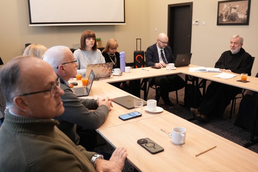 Szeroki kadr z sali konferencyjnej: uczestnicy spotkania siedzą przy stołach ułożonych w podkowę, część pracuje na laptopach, część słucha. W tle po prawej siedzi zakonnik w czarnym habicie, a obok uczestnik w garniturze przy laptopie; na stole widoczne dokumenty, kubki, szklanki i telefony. W centrum sali ekran projekcyjny, a na ścianach czarno-białe zdjęcia w ramkach; atmosfera formalnego zebrania i dyskusji