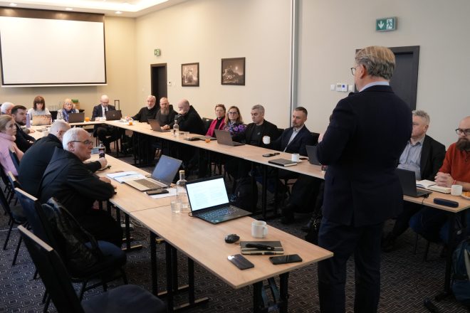 Szeroki kadr sali konferencyjnej: uczestnicy spotkania siedzą po obu stronach stołów ustawionych w podkowę, skupieni na wystąpieniu prowadzącego stojącego tyłem do aparatu. Na stołach rozłożone są laptopy, notatniki, telefony i napoje. Na ścianach wiszą czarno-białe fotografie, a nad stołami znajduje się ekran projekcyjny