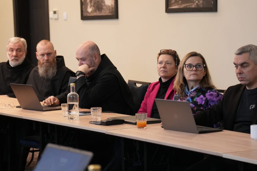 Zbliżenie na rząd uczestników spotkania siedzących przy stole: dwóch zakonników w czarnych habitach oraz osoby świeckie, kobiety i mężczyźni, pracujący na laptopach. Na stole stoją szklanki z wodą i sokiem, butelka oraz notatniki. Uczestnicy patrzą w kierunku prowadzącego, z wyrazem skupienia