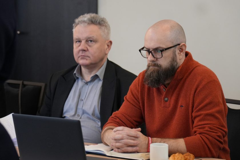 Dwóch mężczyzn siedzących obok siebie przy stole konferencyjnym: jeden w marynarce i koszuli, drugi w czerwonym swetrze i okularach, z długą brodą. Przed nimi otwarty laptop i notes; mężczyzna w czerwonym swetrze ma splecione dłonie i słucha z uwagą. Na stole stoi kubek i talerzyk z rogalikiem