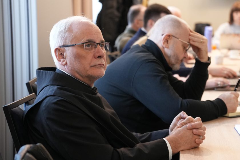 Zbliżenie na zakonnika w czarnym habicie siedzącego przy stole; patrzy w stronę prowadzącego z uważnym, skupionym wyrazem twarzy. Obok niego inny uczestnik pochylony nad notatkami, z dłonią opartą na czole. Na stole widoczne zeszyty i długopis