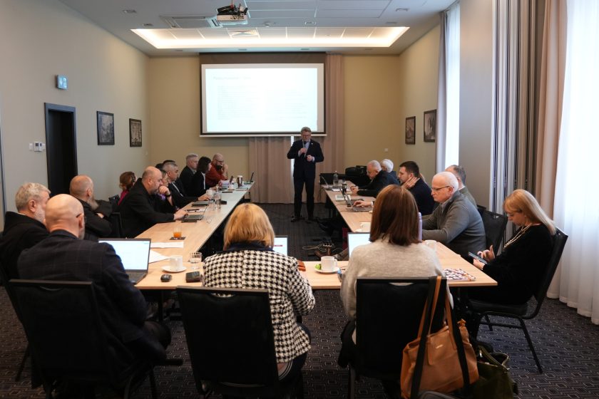 Szeroki widok sali konferencyjnej: prowadzący stoi na środku przed ekranem projekcyjnym, na którym wyświetlony jest slajd z podsumowaniem. Uczestnicy siedzą wokół stołów, część notuje, część pracuje na laptopach. Na stołach znajdują się filiżanki, szklanki, butelki z wodą i dokumenty; pomieszczenie oświetlone jest światłem dziennym wpadającym przez okna z zasłonami