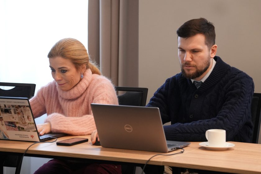 Dwoje uczestników spotkania siedzących przy stole: kobieta w jasnoróżowym swetrze pracuje na laptopie, obok niej mężczyzna w ciemnym swetrze również korzysta z komputera. Na stole leży telefon oraz filiżanka z kawą; oboje są skupieni na ekranach