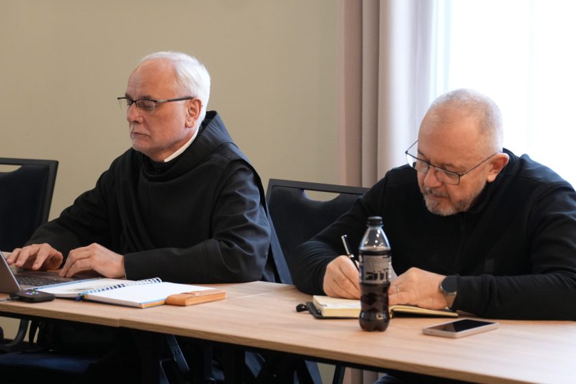 Dwóch zakonników siedzących przy stole konferencyjnym: jeden pracuje na laptopie, drugi zapisuje notatki w zeszycie długopisem. Na stole znajduje się butelka napoju, notes oraz telefon. Obaj mają pochylone głowy i skupioną postawę, charakterystyczną dla pracy roboczej
