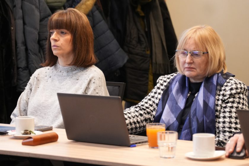 Dwie kobiety siedzące przy stole konferencyjnym, pracujące na laptopie. Jedna z nich ma krótką fryzurę i jasny sweter, druga nosi okulary oraz wzorzysty żakiet i szal. Na stole stoją szklanki z wodą i sokiem oraz filiżanka; w tle widoczne kurtki zawieszone na wieszakach. Obie kobiety patrzą w stronę prowadzącego lub ekranu, słuchając prezentacji