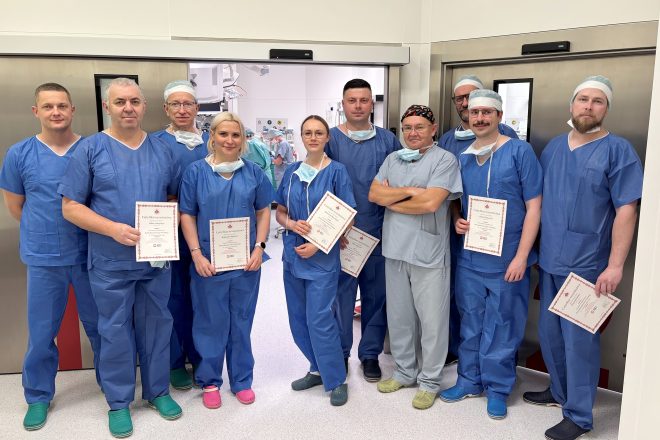 Grupa lekarzy i personelu medycznego ubranych w niebieskie stroje operacyjne stoi w drzwiach bloku operacyjnego. Kilka osób trzyma w dłoniach certyfikaty potwierdzające udział w warsztatach. W tle widoczne jest wnętrze nowoczesnej sali operacyjnej z aparaturą medyczną. Zdjęcie przedstawia uczestników warsztatów leczenia przepuklin zorganizowanych w Szpitalu Zakonu Bonifratrów w Krakowie