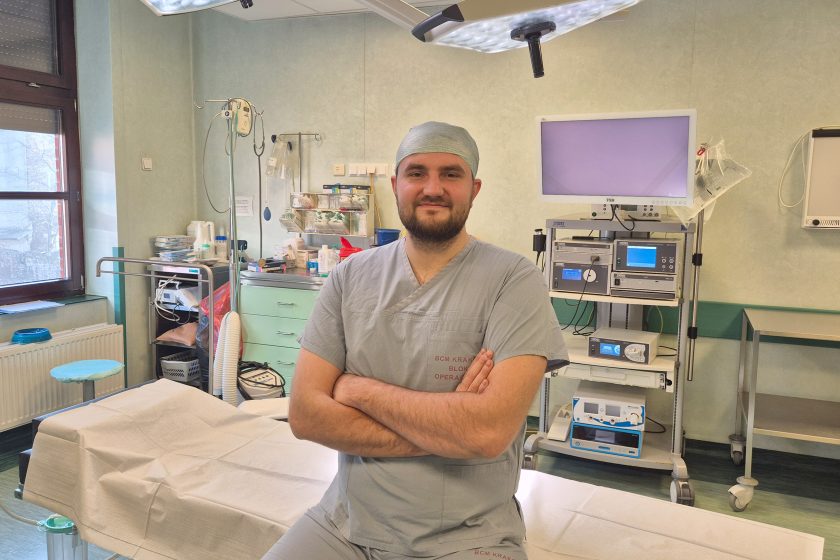 Mężczyzna w stroju operacyjnym siedzi na stole zabiegowym w sali operacyjnej, z założonym czepkiem i skrzyżowanymi rękami. Ma na sobie jasnoszare ubranie medyczne z oznaczeniem bloku operacyjnego. Za nim widoczne są specjalistyczne urządzenia medyczne, w tym monitor, aparatura endoskopowa oraz wózek z narzędziami. Nad stołem znajduje się lampa operacyjna; wnętrze jest jasne i sterylne