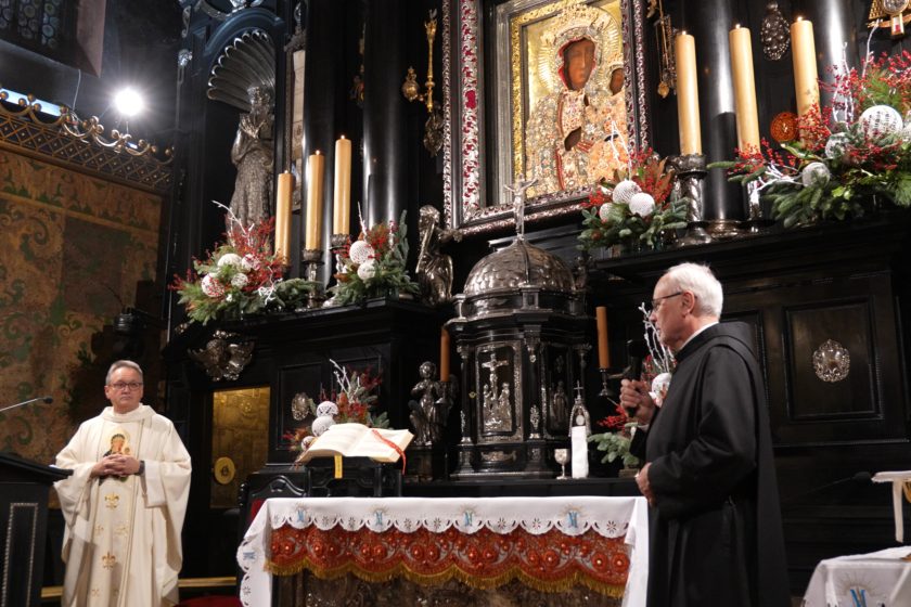 Wnętrze kaplicy na Jasnej Górze: bogato zdobiony, czarny ołtarz z licznymi świecami, srebrnymi elementami i dekoracjami roślinnymi z czerwonymi akcentami. W centralnej części widoczny obraz Matki Bożej Częstochowskiej w ozdobnej ramie. Po lewej stronie stoi kapłan w jasnych szatach liturgicznych, z rękami złożonymi przed sobą; po prawej zakonnik w czarnym habicie przemawia do mikrofonu. Na ołtarzu leży otwarta księga liturgiczna