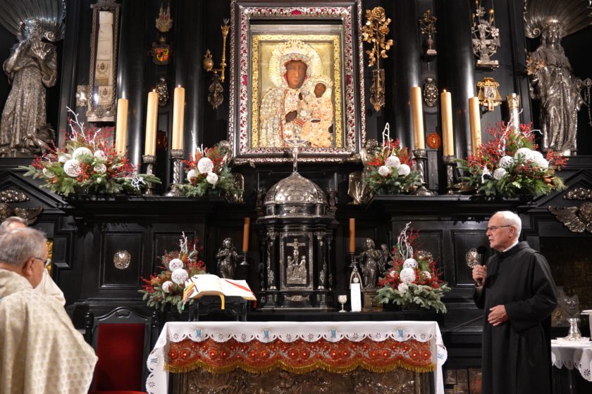 Szeroki kadr wnętrza kaplicy na Jasnej Górze z centralnie umieszczonym obrazem Matki Bożej Częstochowskiej w ozdobnej, srebrno-złotej ramie. Ołtarz wykonany z ciemnego drewna, bogato zdobiony rzeźbami i metaloplastyką; po obu stronach stoją wysokie świece oraz dekoracje roślinne z zieleni i czerwonych akcentów. Na ołtarzu znajduje się otwarta księga liturgiczna. Po prawej stronie kadru zakonnik w czarnym habicie przemawia do mikrofonu, zwrócony w stronę zgromadzonych; po lewej widoczny fragment kapłana w jasnych szatach liturgicznych, stojącego tyłem. Całość oddaje uroczysty, modlitewny charakter nabożeństwa