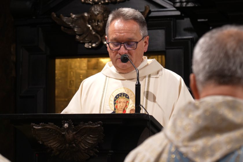 Zbliżenie na kapłana w jasnych szatach liturgicznych stojącego przy ambonie i czytającego tekst do mikrofonu. Na ornacie widoczny wizerunek Matki Bożej. Kapłan ma opuszczony wzrok, skupiony na czytaniu. W tle ciemne, bogato zdobione elementy ołtarza, w tym złocony fragment oraz rzeźbione detale; na pierwszym planie niewyraźne sylwetki innych uczestników liturgii