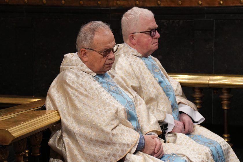 Dwóch kapłanów w jasnych szatach liturgicznych siedzi obok siebie w ławce, w skupieniu uczestnicząc w nabożeństwie