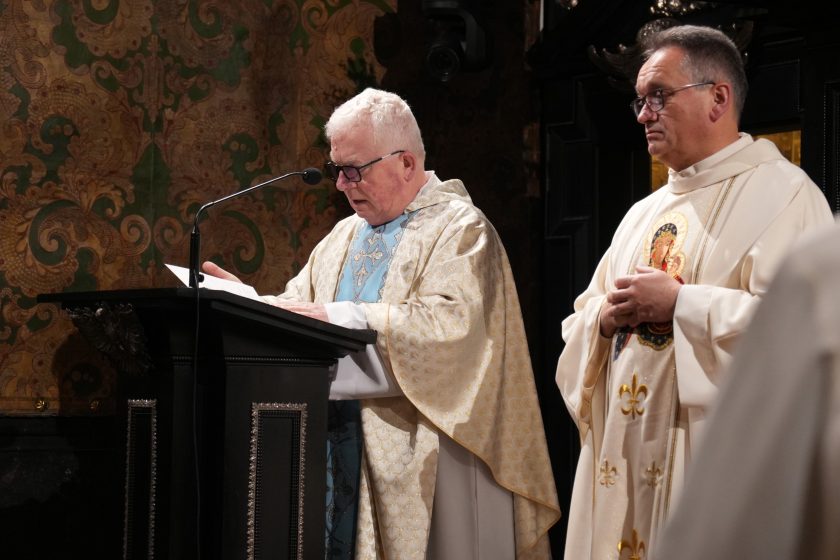 Kapłan w jasnych szatach liturgicznych czyta tekst przy pulpicie, obok stoi drugi duchowny w białym ornacie. Tło stanowi zabytkowe wnętrze kaplicy