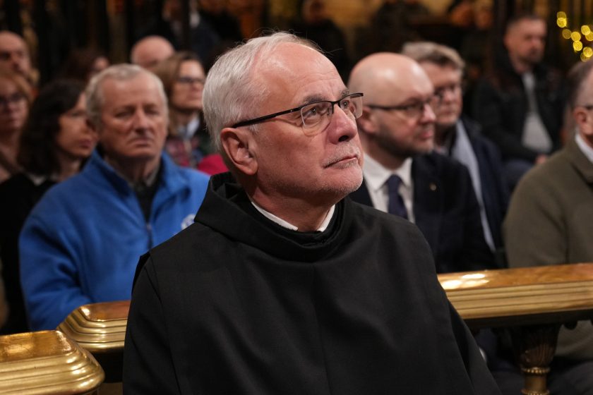 Zbliżenie na zakonnika w czarnym habicie siedzącego w ławce kaplicy, w tle widoczni inni uczestnicy nabożeństwa