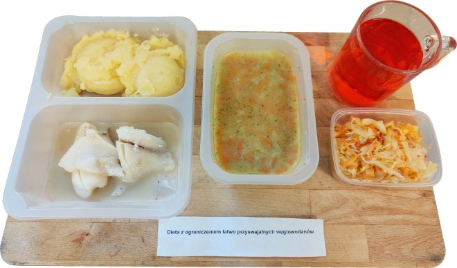 Zestaw obiadowy na diecie z ograniczeniem łatwo przyswajalnych węglowodanów: filet z ryby w jasnym sosie, puree ziemniaczane, zupa jarzynowa, surówka z kapusty oraz herbata owocowa, podane w pojemnikach cateringowych na drewnianej desce