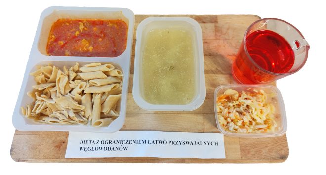 Obiad szpitalny w ramach diety z ograniczeniem łatwo przyswajalnych węglowodanów: makaron pełnoziarnisty z sosem pomidorowym z dodatkiem mięsa, zupa klarowna, surówka z kapusty oraz herbata owocowa. Posiłki podane w pojemnikach jednorazowych na drewnianej desce
