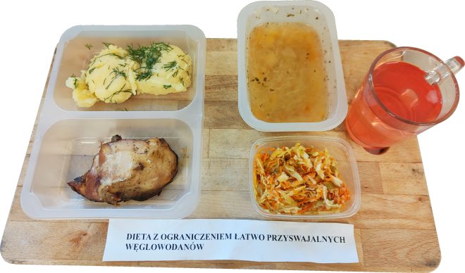 Zestaw obiadowy w diecie z ograniczeniem łatwo przyswajalnych węglowodanów: pieczone mięso, puree ziemniaczane z koperkiem, surówka z kapusty i marchwi, zupa jarzynowa oraz napój, podane w pojemnikach na drewnianej desce