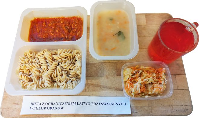 Zestaw obiadowy w diecie z ograniczeniem łatwo przyswajalnych węglowodanów: makaron świderki z sosem mięsnym, zupa jarzynowa, surówka z kapusty i marchewki oraz kompot owocowy, podane w jednorazowych pojemnikach na drewnianej desce