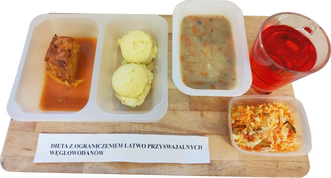 Na jasnej drewnianej desce stoi zestaw posiłków w białych pojemnikach. Po lewej w prostokątnym pojemniku widać kawałek pieczonego mięsa w czerwonym sosie. Obok znajduje się osobny pojemnik z dwiema porcjami puree ziemniaczanego. Dalej ustawiony jest pojemnik z jasną zupą z widocznymi kawałkami marchewki i ziół. W prawym dolnym rogu leży mały pojemnik z surówką z drobno posiekanej kapusty i marchewki. Po prawej stronie stoi szklany kubek z czerwonym napojem. Na dole zdjęcia widoczna kartka z napisem: „DIETA Z OGRANICZENIEM ŁATWO PRZYSWAJALNYCH WĘGLOWODANÓW”