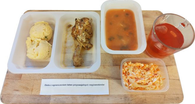 Zestaw obiadowy na drewnianej desce, w pojemnikach: po lewej dwa kopki purée ziemniaczanego, obok udko z kurczaka; po prawej pojemnik z czerwoną zupą (np. pomidorową) oraz szklany kubek z czerwonym napojem. Na pierwszym planie mały pojemnik z surówką z kapusty i marchewki; na dole kartka z napisem „Dieta z ograniczeniem łatwo przyswajalnych węglowodanów”