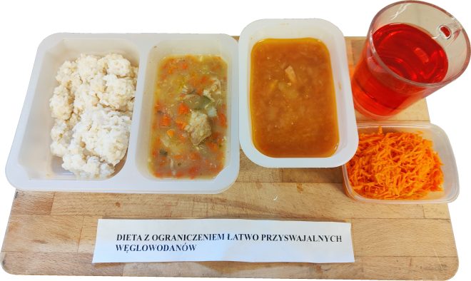 Obiad w diecie z ograniczeniem łatwo przyswajalnych węglowodanów: w pojemniku porcje białego ryżu oraz gulasz mięsny z warzywami w sosie; obok osobny pojemnik z brązowym sosem, surówka z tartej marchewki oraz szklanka czerwonego napoju. Posiłek ustawiony na drewnianej desce