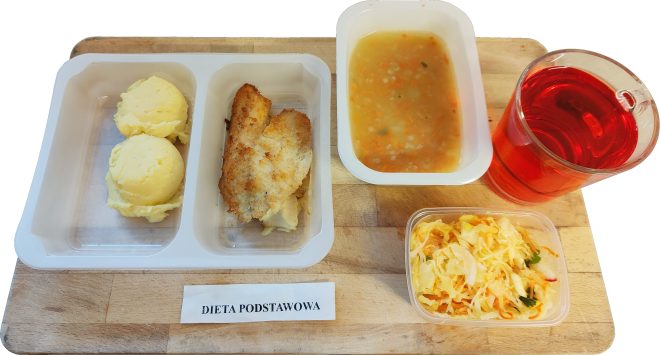 Obiad w diecie podstawowej podany na drewnianej desce. W plastikowej tacce znajdują się dwie porcje purée ziemniaczanego oraz filet rybny panierowany i smażony. Obok ustawiona jest miseczka z jarzynową zupą oraz szklanka czerwonego napoju. Dodatkowo widoczna jest surówka z białej kapusty z marchewką. Na desce umieszczona kartka z napisem „Dieta podstawowa”