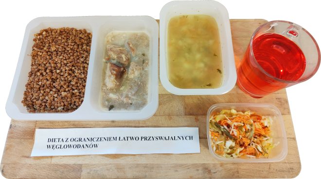 Zestaw obiadowy na diecie z ograniczeniem łatwo przyswajalnych węglowodanów: kasza gryczana, porcja mięsa w sosie, zupa jarzynowa, surówka z kapusty i marchewki oraz napój owocowy, podane na drewnianej desce