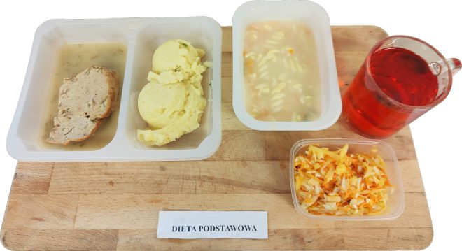 Zestaw obiadowy ułożony na drewnianej desce, opisany jako „Dieta podstawowa”. Po lewej stronie znajduje się plastikowy pojemnik dwudzielny: w jednej części porcja mięsa w jasnym sosie, w drugiej porcja puree ziemniaczanego. Po prawej stronie widoczny osobny pojemnik z zupą jarzynową z drobnym makaronem. Obok stoi szklany kubek z czerwonym napojem. W prawym dolnym rogu znajduje się mały pojemnik z surówką z kapusty i marchwi. Wszystkie elementy są wyraźnie oddzielone i podane w jednorazowych naczyniach