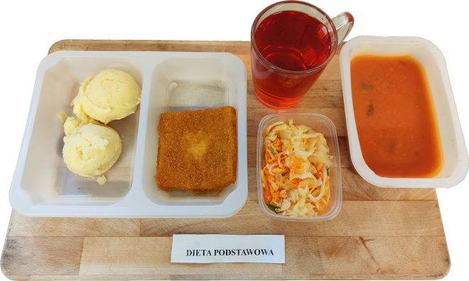 „Na drewnianej desce ułożony zestaw obiadowy w ramach diety podstawowej. Po lewej stronie plastikowa tacka z dwoma porcjami puree ziemniaczanego oraz panierowanym kotletem w kształcie prostokąta. Obok mały, przezroczysty pojemnik z surówką z kapusty i marchwi. Po prawej stronie znajduje się miska z zupą pomidorową oraz szklany kubek z czerwonym napojem. Na dole deski widoczna etykieta z napisem: ‘Dieta podstawowa’
