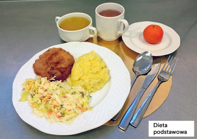 Zestaw obiadowy w diecie podstawowej podany na metalowym blacie. Na białym talerzu znajdują się: kotlet w panierce, porcja purée ziemniaczanego oraz surówka z białej kapusty z dodatkiem marchewki i zieleniny. W tle ustawiono miseczkę z jasną zupą oraz kubek z herbatą. Obok, na małym talerzyku, leży mandarynka. Przy talerzu ułożone są sztućce na okrągłej, złotej podkładce; w prawym dolnym rogu widoczny napis „Dieta podstawowa”
