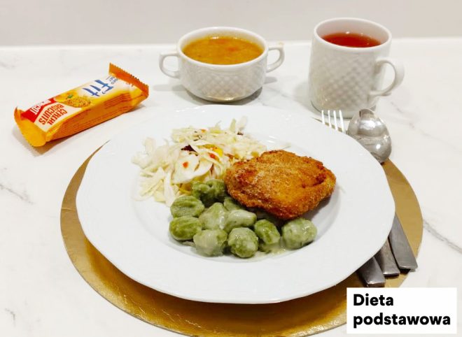 Obiad w diecie podstawowej podany na jasnym blacie. Na białym talerzu znajduje się kotlet w panierce, porcja zielonych klusek podanych z jasnym sosem oraz surówka z białej kapusty. W tle stoją: miseczka z zupą oraz kubek z herbatą. Po lewej stronie kadru widoczny jest zapakowany słodki dodatek (wafelek). Przy talerzu ułożone są sztućce na okrągłej, złotej podkładce; w prawym dolnym rogu znajduje się napis „Dieta podstawowa”