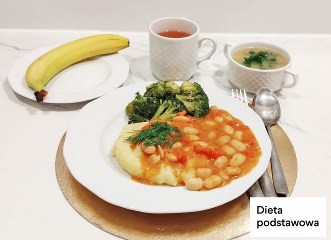Obiad w diecie podstawowej: fasolka w sosie pomidorowym podana z puree ziemniaczanym, gotowanymi brokułami i koperkiem. Obok talerza banan, zupa jarzynowa oraz kubek z herbatą. Na zdjęciu widoczny napis „Dieta podstawowa”