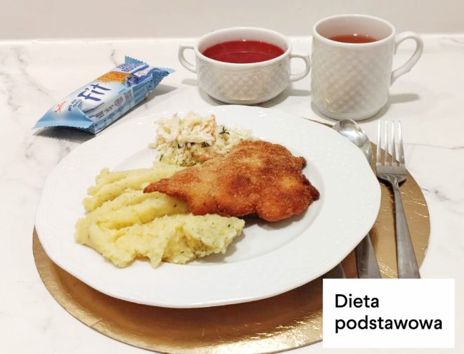 Obiad w diecie podstawowej: panierowany kotlet smażony podany z puree ziemniaczanym i surówką z kapusty. Obok talerza miseczka z czerwonym barszczem oraz kubek z herbatą. Na zdjęciu widoczny napis „Dieta podstawowa” i herbatniki w opakowaniu z napisem FIT