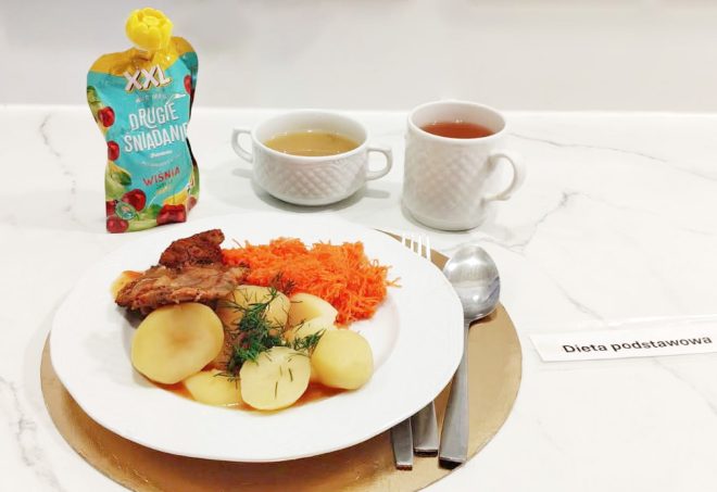 Obiad w ramach diety podstawowej: gotowane ziemniaki z sosem, porcja pieczonego mięsa oraz surówka z tartej marchewki. Zestaw uzupełniają zupa, herbata oraz napój owocowy podany w opakowaniu