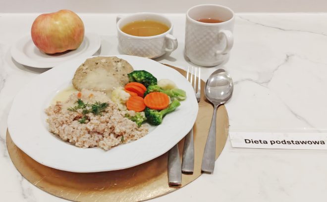Obiad w diecie podstawowej: kasza jęczmienna z sosem koperkowym, kotlet mielony w sosie, gotowane warzywa – brokuł, marchew i kalafior. Obok miseczka zupy, kubek herbaty oraz jabłko na talerzyku. Widoczna etykieta „Dieta podstawowa”