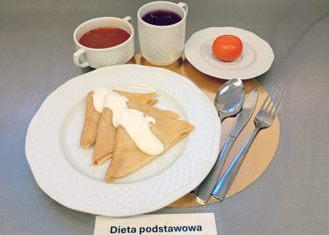 Obiad w diecie podstawowej: naleśniki podane z jasnym sosem na wierzchu. W tle miseczka czerwonej zupy, kubek herbaty oraz mandarynka na talerzyku. Na dole kadru oznaczenie „Dieta podstawowa”