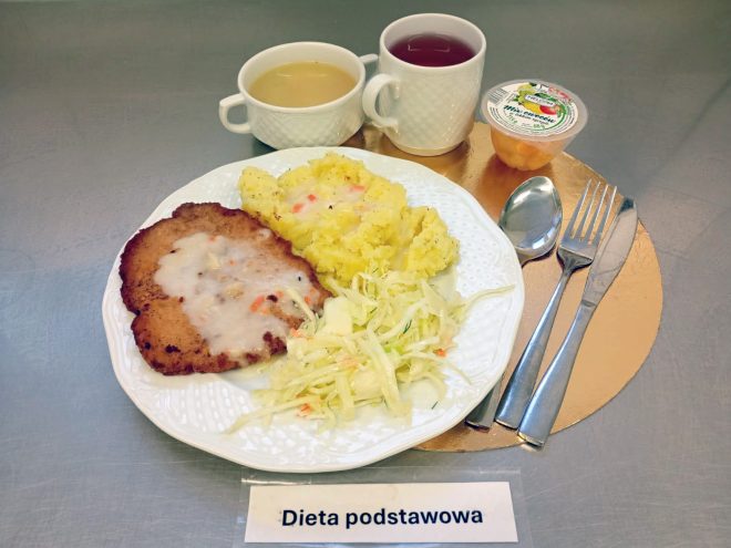 Obiad w „Diecie podstawowej” ułożony na okrągłej, złotej podkładce na metalowym blacie. Na białym talerzu znajduje się kotlet w jasnym sosie, porcja tłuczonych ziemniaków oraz surówka z białej kapusty. Z tyłu stoją dwa naczynia: miseczka z jasną zupą i kubek z czerwonym napojem. Po prawej leżą sztućce (łyżka, widelec i nóż), a z tyłu po prawej znajduje się zamknięty kubeczek z owocami/deserem