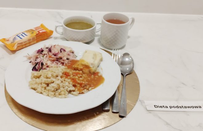 Obiad w „Diecie podstawowej” na marmurowym blacie, na okrągłej złotej podkładce. Na białym talerzu znajduje się porcja kaszy oraz kawałek dania w sosie (jasny filet lub zapiekanka polana sosem), a obok surówka z jasnej i fioletowej kapusty. Z tyłu stoją dwa naczynia: miseczka z jasną zupą i kubek z herbatą. Po lewej widać zapakowany wafelek/deser, a po prawej ułożone sztućce (łyżka i widelec) oraz etykietę „Dieta podstawowa”