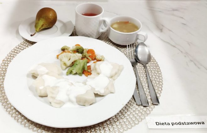 Na jasnym, marmurowym blacie stoi zestaw obiadowy opisany jako „Dieta podstawowa”. Na dużym białym talerzu znajdują się jasne pierogi lub kluski polane białym sosem oraz porcja gotowanych warzyw (brokuł, kalafior i marchew). U góry kadru widać kubek z czerwonym napojem oraz małą miseczkę z jasną zupą. Po lewej na małym talerzyku leży gruszka. Po prawej ułożone są sztućce: widelec, łyżka i nóż