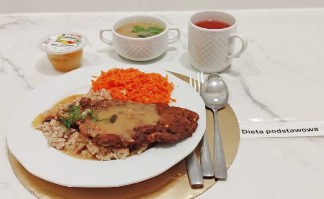 Na jasnym, marmurkowym blacie podano zestaw obiadowy „Dieta podstawowa”: na dużym białym talerzu kotlet/porcja mięsa w jasnym sosie z dodatkiem kaszy oraz surówka z tartej marchewki. Obok leżą sztućce. W tle stoją: miseczka z jasną zupą z zieleniną, kubek z czerwonym napojem oraz mały kubeczek z deserem/owocowym musem; z boku widoczna kartka z napisem „Dieta podstawowa”